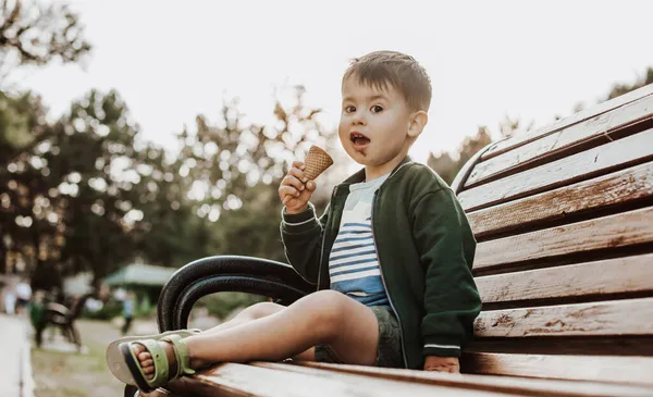 Cucaian menino comendo sorvete sentado em um banco de parque, olhando para a câmera — Fotografia de Stock