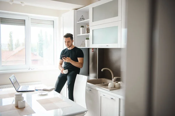 A man in the kitchen while having breakfast sends a message or calls with the phone. Laptop on the table. Working from home. Concept of: social network. — Stock Photo, Image