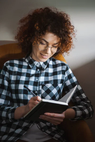 Retrato de una mujer caucásica sentada en un sillón, escribiendo en un planificador. Cabello rizado. — Foto de Stock