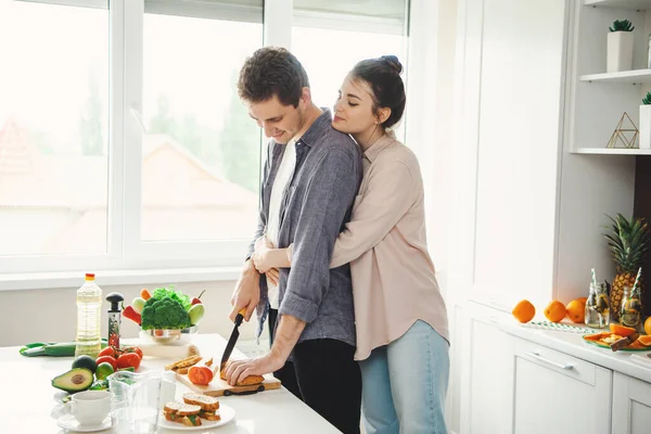 Giovane ragazza che abbraccia il suo ragazzo mentre taglia il pane in cucina. Cibo sano vegetariano. Cucinare insieme. Uno stile di vita sano. Cucina familiare. — Foto Stock