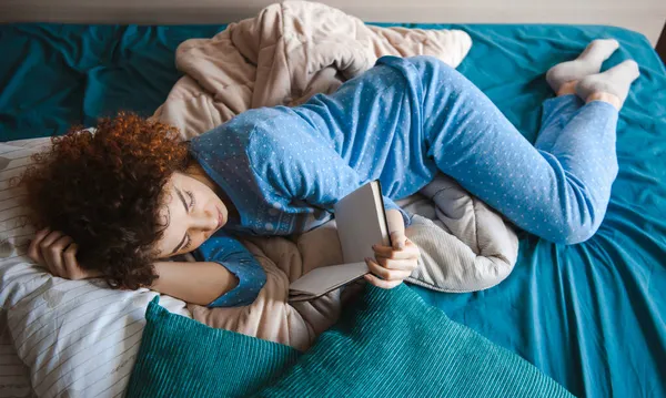 Λευκή γυναίκα διαβάζει βιβλίο το πρωί στο σπίτι, στο σαλόνι, ξαπλωμένη στο κρεβάτι με κοιμισμένα ρούχα. Ώρα για ύπνο. Σγουρά μαλλιά — Φωτογραφία Αρχείου