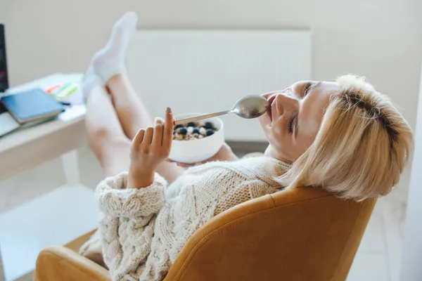 Vista posterior de una mujer caucásica comiendo sabrosa avena dulce en casa sentada relajada en el sillón. Alimentos orgánicos naturales. — Foto de Stock
