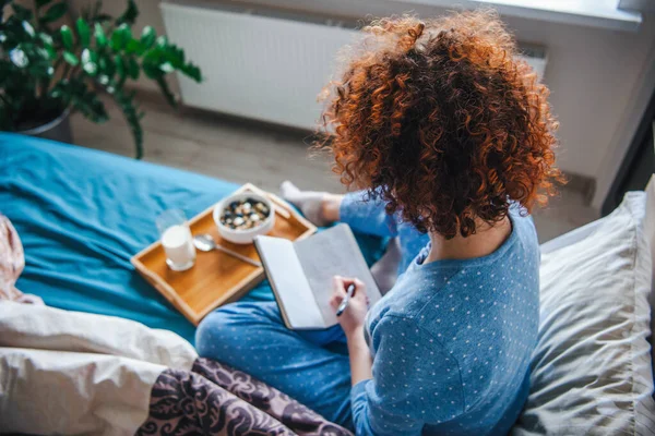 Vista trasera de una mujer rizada sentada en la cama notando algo en su cuaderno. Bandeja con sabroso desayuno en la cama. — Foto de Stock