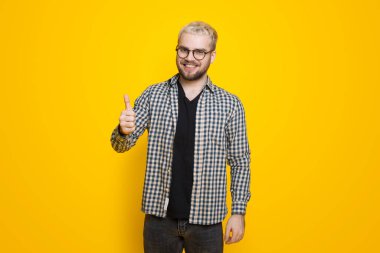 Sakallı, gözlüklü beyaz bir adam sarı arka planda el izole edilmiş mutlu baş parmaklar gösteriyor. İşaret gibi