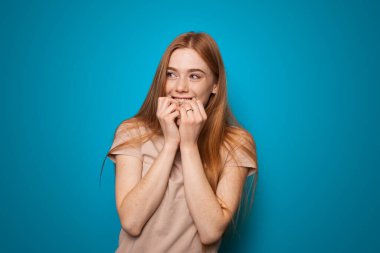 Heyecanlı kızıl saçlı kadın çenesine dokunuyor ve gülümsüyor mavi arka planda iyi haberleri beklerken.