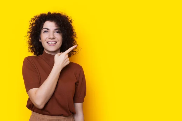 Framifrån av en lycklig kvinna står isolerad över gul bakgrund tittar på kameran och pekar med pekfingret på emty utrymme. Kopieringsutrymme. Lockigt hår — Stockfoto