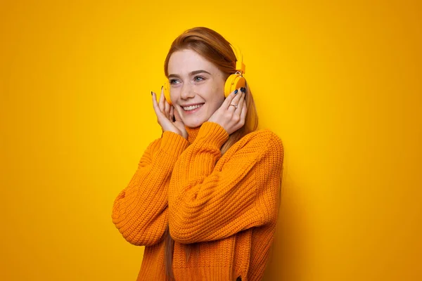 Portrét usměvavé zrzavé ženy, která poslouchá hudbu na sluchátkách ve studiu na žlutém pozadí. — Stock fotografie
