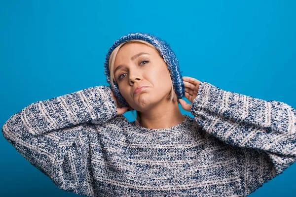 Retrato de primer plano de mujer rubia triste con emociones sinceras tocando la cabeza usando suéter azul cálido y sombrero de punto azul aislado sobre fondo azul. — Foto de Stock
