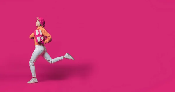 Vista lateral de longitud completa de la mujer corriendo sosteniendo la caja presente mirando hacia otro lado aislado sobre fondo de estudio rosa. Copiar espacio. —  Fotos de Stock