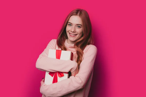 Mujer de ensueño abrazando su regalo sorpresa y sonriendo mientras mira a la cámara, de pie sobre fondo rosa. Pelirroja mujer caucásica. Hermosa chica. Regalo de Navidad. — Foto de Stock