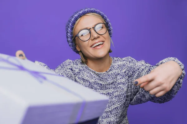 Mujer caucásica celebrando la Navidad, sosteniendo un regalo apuntando con el dedo índice para presentar la caja aislada sobre fondo púrpura. Concepto de celebración de cumpleaños — Foto de Stock