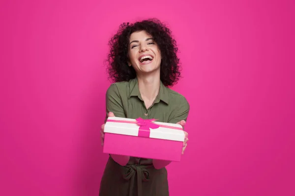 Mujer rizada sonriente en vestido posando mientras sostiene la caja de regalo con lazo de cinta de regalo aislado sobre fondo rosa. Sorpresa de regalo navideño. Cumpleaños actual. — Foto de Stock