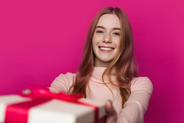 Mujer con pelirroja y pecas dando caja de regalo y sonriendo aislada sobre fondo rosa. Felicitaciones por cumpleaños y regalo de ofrenda. Cara pecosa. Navidad. — Foto de Stock