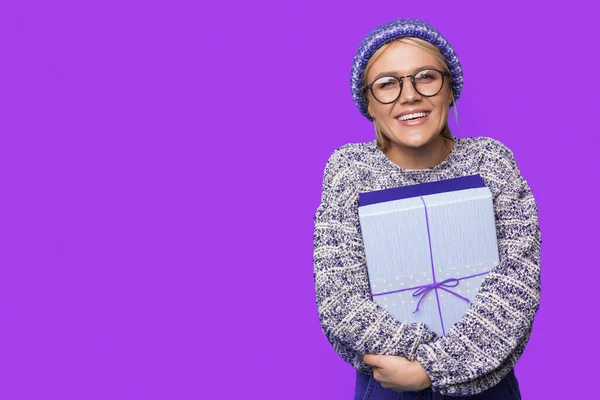 Widok z przodu kobiety trzymającej obecne pudełko świętujące Nowy Rok w swetrze z dzianiny i kapeluszu, pozujących na fioletowym tle z przestrzenią do kopiowania. Patrzę w kamerę. — Zdjęcie stockowe