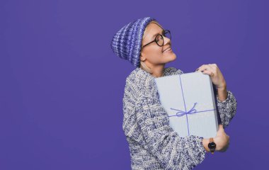 Beyaz bir kadının örgü süveter giyen bir hediye kutusunu kucaklarken çekilmiş yan görüntüsü. Mor arkaplandan izole edilmiş, kopyalama alanı olan bir süveter..