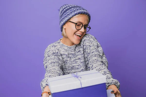 Mujer caucásica sosteniendo una caja de regalo hacia la cámara mientras sonríe con los ojos cerrados aislados sobre fondo púrpura. Retrato de niña bonita feliz sonriente — Foto de Stock