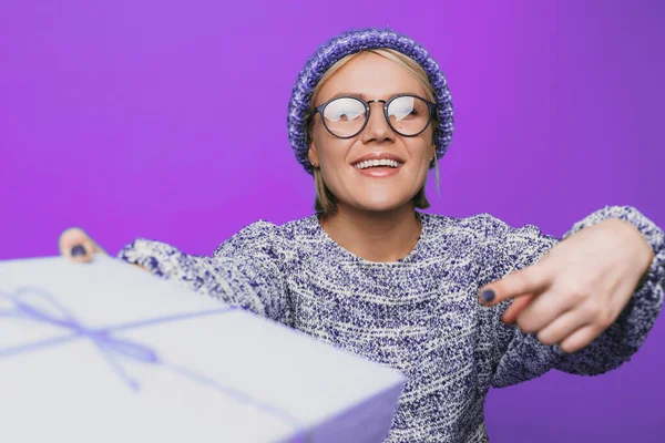 Mujer sonriente en suéter cineteado que sostiene el regalo y el dedo índice a la caja enfondo aislado púrpura. Usando anteojos. Caja de regalo de Navidad. Navidad. — Foto de Stock