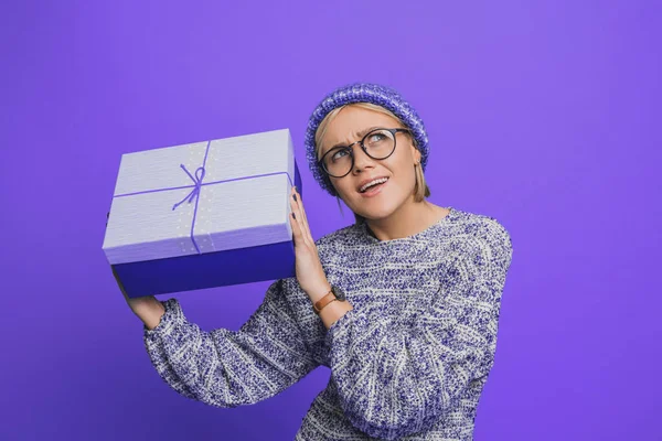 Curiosa mujer caucásica con suéter de punto sacudiendo caja de regalo tratando de adivinar lo que está dentro aislado sobre fondo púrpura. Sorpresa de regalo navideño. Navidad. — Foto de Stock