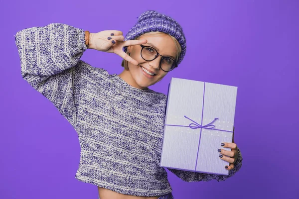 Retrato de una mujer con sombrero de punto sosteniendo caja de regalo que se divierte aislado sobre fondo de color púrpura. Mostrando signo v. Concepto de vacaciones de invierno y primavera — Foto de Stock
