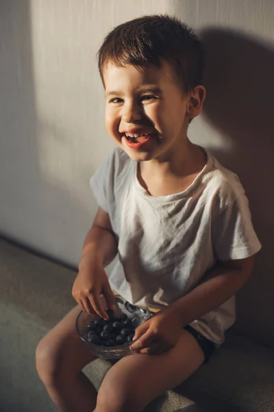 Garoto branco comendo mirtilos frescos de uma tigela sorrindo e sentado no sofá enquanto olha para a câmera. — Fotografia de Stock