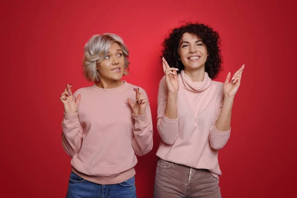 Photo de deux femmes tenant les doigts croisés souhaitant bonne chance isolées sur fond de couleur rouge. Cheveux bouclés — Photo