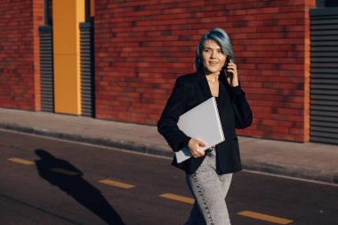 Mavi saçlı, beyaz bir kadın elinde dizüstü bilgisayarla telefon görüşmesi yaparken dışarı bakıyor. Çalışma gününün sonunda