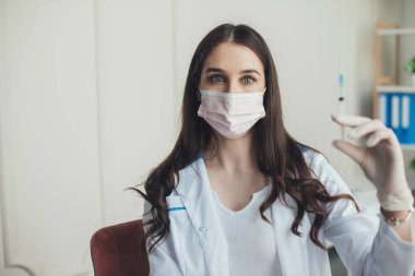 Aşı için şırıngayla poz veren hemşire beyaz bornoz, lastik eldiven ve tıbbi maske takıyor. Corona virüsü aşılama. Tıbbi tedavi.