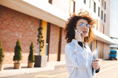 Kollarında dizüstü bilgisayarla telefonda konuşan bir iş kadınının yan görüntüsü. Kıvırcık saçlı kadın