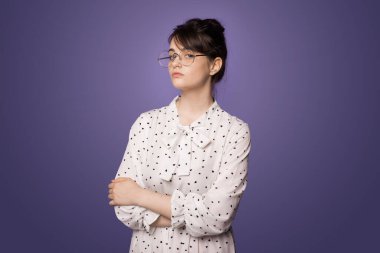 Kollarını kavuşturmuş, kameraya şüpheci bir ifadeyle bakan bir kadının portresi. Gözlük takıyordu. Mor arkaplan üzerinde izole edilmiş