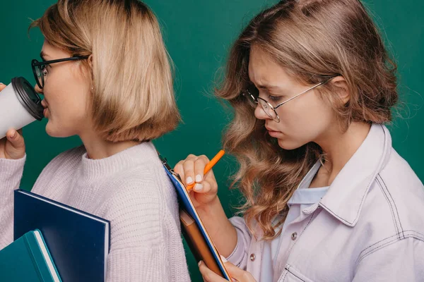 Ein Mädchen trinkt Kaffee, während das andere ihr Notizbuch hinter ihre Freundin lehnt, damit sie Hausaufgaben schreiben kann. Seitenansicht von zwei Frauen isoliert auf grünem Hintergrund. — Stockfoto