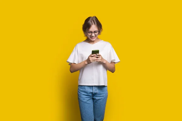 Běloška stojí izolovaně na žlutém pozadí a dívá se na obrazovku telefonu, zatímco píše meesage. Usměvavá tvář — Stock fotografie