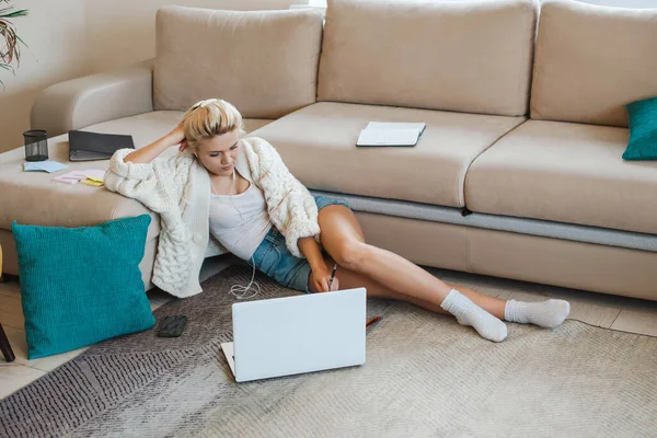 Mujer caucásica usando su computadora portátil mientras está acostada en el suelo y apoyada en el sofá. Estudiando desde casa. Educación a distancia. — Foto de Stock