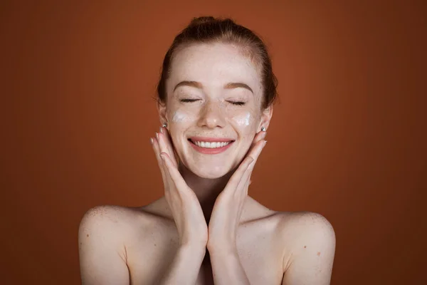 Blanke sproeten vrouw aanbrengen cosmetische crème op haar gezicht geïsoleerd over bruine achtergrond. Glimlachende roodharige vrouw. — Stockfoto