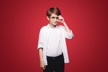 Gözlük tutan konsantre bir çocuğun ön görüntüsü ve kırmızı bir arkaplanda izole edilmiş kameraya bakıyor. Okul çocukları