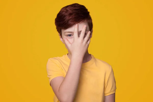 Retrato de um garoto vestindo camiseta casual em pé sobre fundo amarelo isolado cobrindo parte do rosto com a mão. Gestos. Infância — Fotografia de Stock