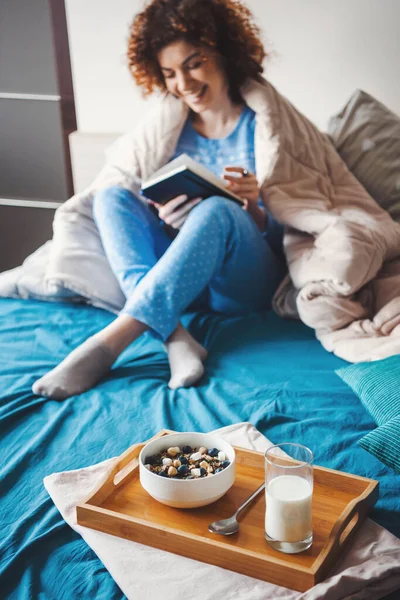 Wanita berambut keriting kaukasia membaca buku di tempat tidur dan makan sereal dengan susu. Makanan sehat. Pagi di tempat tidur — Stok Foto