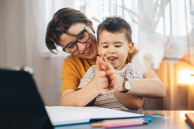 Beyaz bir anne evinde bir masada oturup oğluna ödevlerinde yardım ediyor. Dizüstü bilgisayar kullanarak ona bir şey gösteriyor. Online eğitim. Çalışıyorum, ödev yapıyorum..