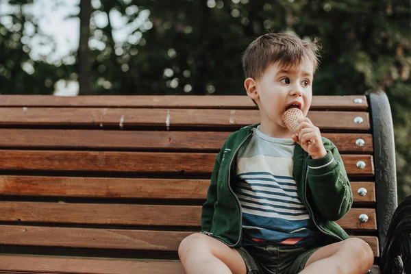 Vista frontal de um menino caucasiano comendo sorvete sentado em um banco no parque olhando para longe. Espaço livre. Infância. — Fotografia de Stock