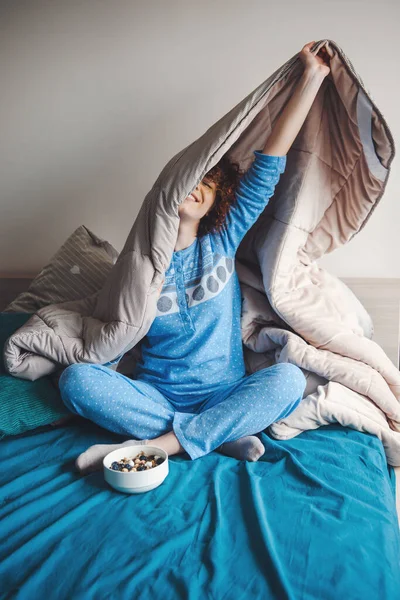 Mujer rizada que pasa su fin de semana en la cama debajo de una manta, vistiendo pijamas azules. Comida saludable. Desayuno en la cama — Foto de Stock