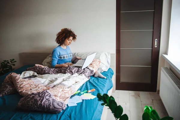 Mujer rizada caucásica haciendo lecciones en línea desde la cama usando un portátil en un pijama azul. Estilo de vida cuarentena. — Foto de Stock