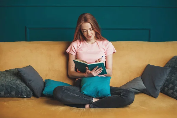 Framifrån av en ingefära kvinna läser bok när du sitter på soffan hemma. Korsade ben. En rödhårig kvinna. Övergångsbestämmelser — Stockfoto
