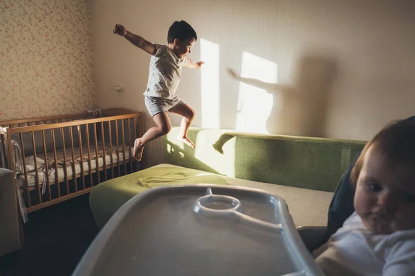 O menino pulando no sofá enquanto o irmão mais novo espera para ser alimentado — Fotografia de Stock