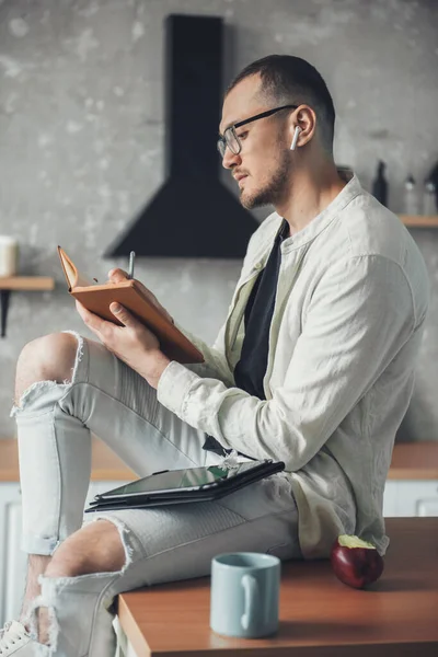 Hombre sentado en la mesa de la cocina con portátil, utilizando la tableta. Trabajando desde casa. Modelo de moda. Estilo de vida cuarentena. —  Fotos de Stock