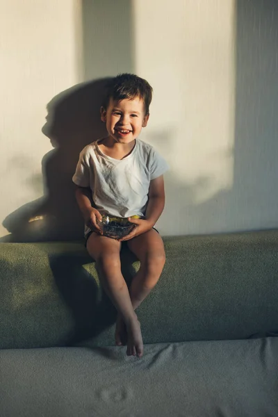 Menino caucasiano sorrindo para a câmera enquanto come uma tigela com mirtilos sentados no sofá em casa. — Fotografia de Stock