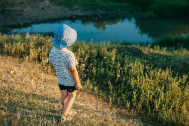 Baraj gölünün kıyısında oturan mavi şapkalı bir çocuk. Parktaki göle bakıyorum..