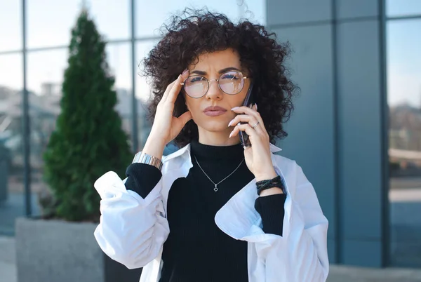 Curly manager frowning concerned speaking on cellphone outside. Close-up portrait of a caucasian woman looking on camera. Mobile phone talk concept — Zdjęcie stockowe