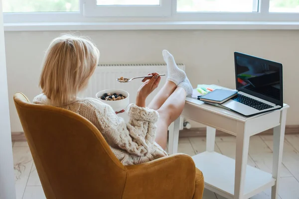 Vista trasera trasera. Mujer que trabaja desde casa en el ordenador portátil mientras come la comida con muesli. Mañana. Estilo de vida saludable. Vista trasera. — Foto de Stock