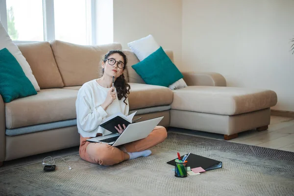 Woman studying at home with crossed legs on the floor leaning against a sofa working with a laptop and class notes. Caucasian woman looking up while thinking — Stock Photo, Image