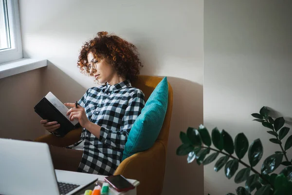 Curly woman in eyeglasses sitting in armchair with laptop on small table aside and taking notes in planner. Freelance career. Business meeting. Business woman — Stockfoto