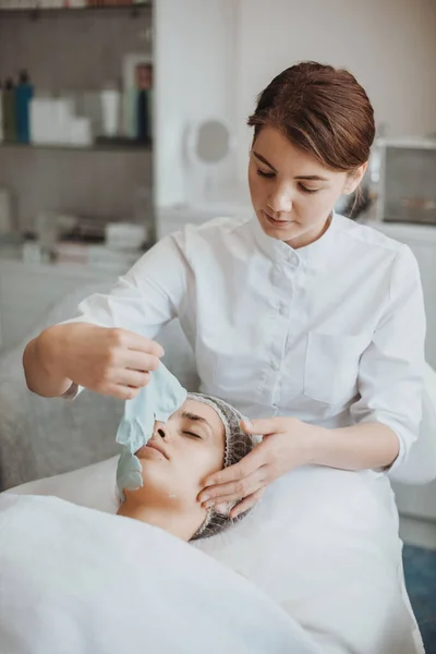 Косметолог видаляє маску з білого крему на обличчі білої жінки, яка лежить на дивані під час чищення обличчя та косметичних процедур . — стокове фото